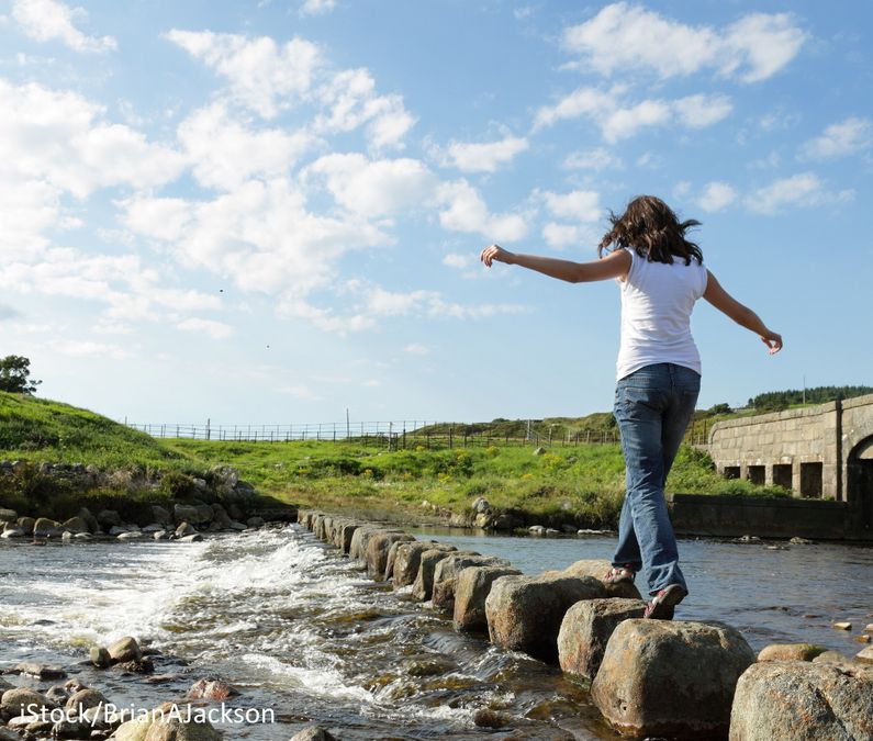 Symbolbild: Eine Frau balanciert auf Steinen über einen Fluss